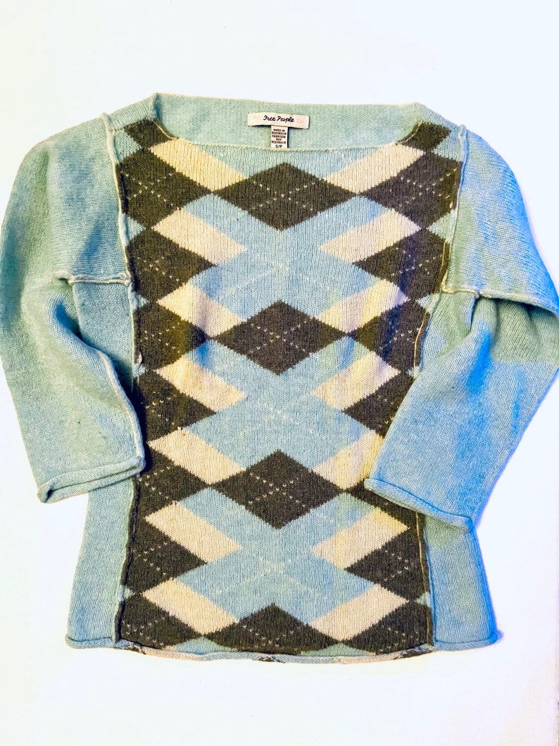 Puede incluir: Un su&eacute;ter azul claro con un patr&oacute;n de diamantes en blanco y marr&oacute;n. El su&eacute;ter tiene un cuello redondo y mangas largas. La etiqueta de la marca Free People es visible en la parte delantera del su&eacute;ter.