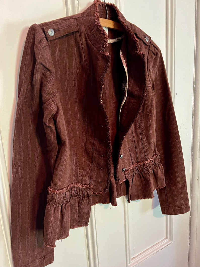 May include: A brown striped jacket with a ruffled hem and frayed edges. The jacket has a stand-up collar and a button closure.