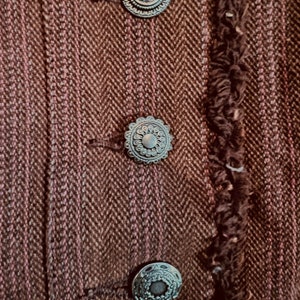 May include: Close-up of a brown tweed jacket with three decorative silver buttons. The buttons have a circular design with a raised center and intricate details.