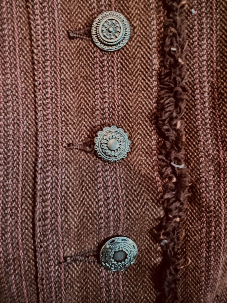 May include: Close-up of a brown tweed jacket with three decorative silver buttons. The buttons have a circular design with a raised center and intricate details.