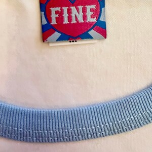 May include: A white t-shirt with a light blue ribbed trim around the neck and sleeves. The shirt has a small, red heart-shaped tag with the word "FINE" printed in white letters.