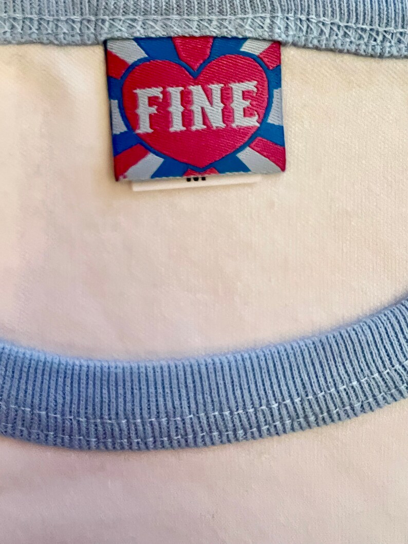 May include: A white t-shirt with a light blue ribbed trim around the neck and sleeves. The shirt has a small, red heart-shaped tag with the word "FINE" printed in white letters.