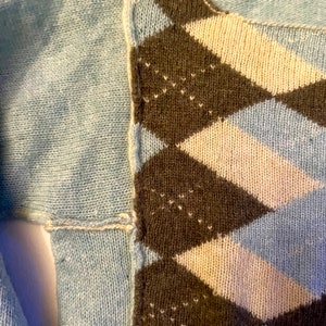 Puede incluir: Primer plano de un su&eacute;ter azul con un patr&oacute;n de rombos marr&oacute;n, blanco y azul. El su&eacute;ter est&aacute; tejido y tiene una costura en el lateral.