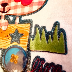 May include: A white fabric patch with a pink and white gingham design, a red flower with a black centre, a blue star, a yellow and brown wooden box, green grass, a blue and gold ball, and a red and blue sparkly word "fun".
