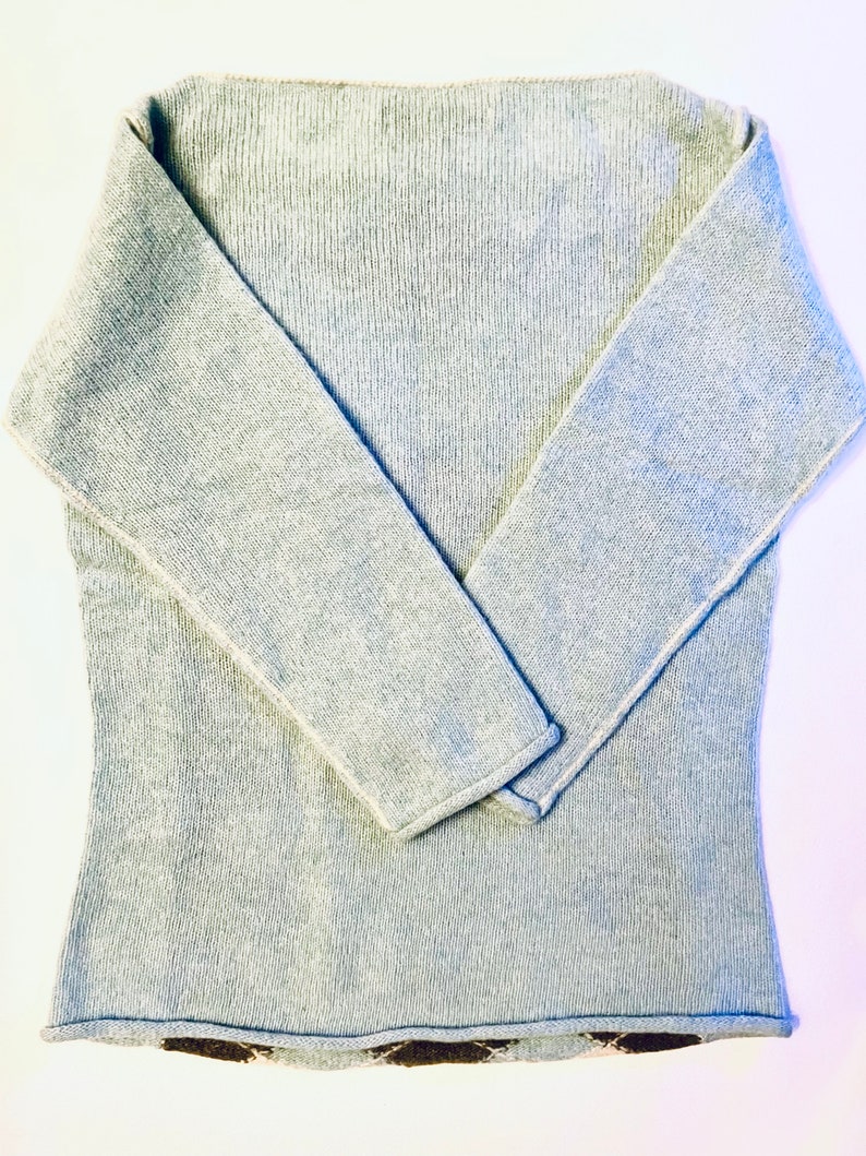 Puede incluir: Un su&eacute;ter gris claro con mangas largas y textura acanalada. El su&eacute;ter tiene un ribete contrastante marr&oacute;n y blanco alrededor del borde inferior.