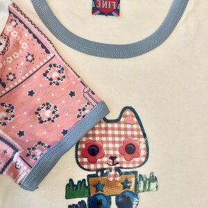 May include: White t-shirt with a blue trim around the neck and sleeves. The shirt features a pink bandana print on one sleeve and a cartoon cat wearing a red and white checked dress and holding a wooden box with stars on it.