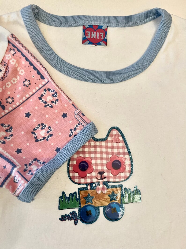 May include: White t-shirt with a blue trim around the neck and sleeves. The shirt features a pink bandana print on one sleeve and a cartoon cat wearing a red and white checked dress and holding a wooden box with stars on it.