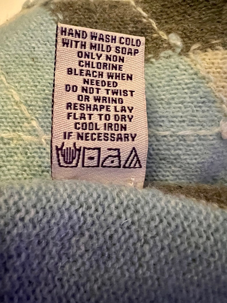 Puede incluir: Una etiqueta de cuidado blanca con texto negro que dice "HAND WASH COLD WITH MILD SOAP ONLY NON CHLORINE BLEACH WHEN NEEDED DO NOT TWIST OR WRING RESHAPE LAY FLAT TO DRY COOL IRON IF NECESSARY". La etiqueta tambi&eacute;n tiene un s&iacute;mbolo en blanco y negro para el lavado a mano.