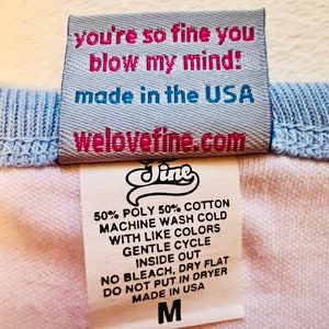 May include: A white and blue clothing label with the text "you're so fine you blow my mind!" and "made in the USA". The label also includes the website "welovefine.com" and the brand name "Jine". The label lists care instructions for the garment, including machine wash cold, gentle cycle, inside out, no bleach, dry flat, do not put in dryer, and made in the USA. The label also indicates the size "M".