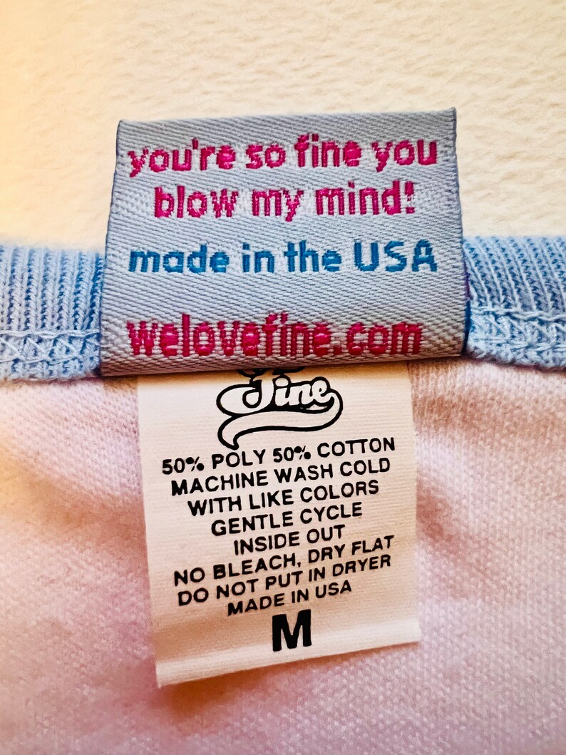May include: A white and blue clothing label with the text "you're so fine you blow my mind!" and "made in the USA". The label also includes the website "welovefine.com" and the brand name "Jine". The label lists care instructions for the garment, including machine wash cold, gentle cycle, inside out, no bleach, dry flat, do not put in dryer, and made in the USA. The label also indicates the size "M".