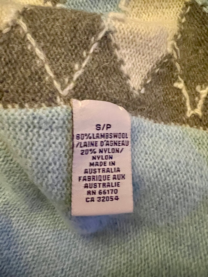 Puede incluir: Una etiqueta blanca con texto negro en un su&eacute;ter azul claro. La etiqueta dice "S/P 80% LAMBSWOOL LAINE D'AGNEAU 20% NYLON/ NYLON MADE IN AUSTRALIA FABRIQUE AUX AUSTRALIE RN 65170 CA 32054".