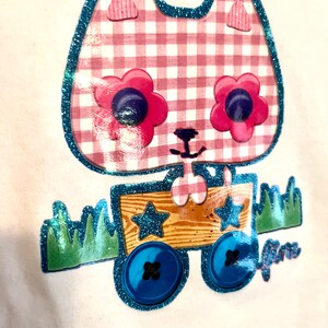 May include: A white t-shirt featuring a pink and white gingham cat design. The cat has blue glitter eyes and is pulling a wooden wagon with blue glitter wheels. The wagon has two stars on the side. The text "Janie" is on the bottom right of the design.