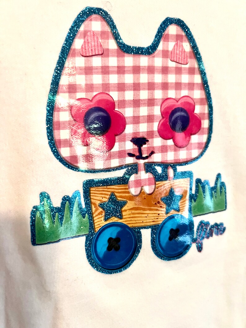 May include: A white t-shirt featuring a pink and white gingham cat design. The cat has blue glitter eyes and is pulling a wooden wagon with blue glitter wheels. The wagon has two stars on the side. The text "Janie" is on the bottom right of the design.
