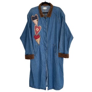 May include: A long, blue denim coat with a brown corduroy collar and cuffs. The coat features three heart-shaped patches with patriotic designs. The coat is hanging on a black hanger.