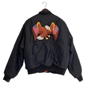 May include: Black bomber jacket with a large embroidered eagle patch on the back. The eagle has brown feathers, a white head, and is holding American flags in its talons. The jacket has a ribbed collar, cuffs, and waistband.