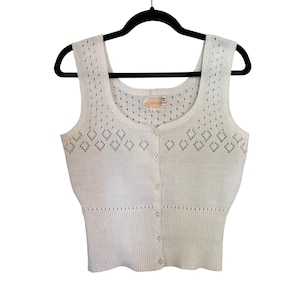 May include: A sleeveless white knit tank top featuring a scoop neckline, a front button-down closure with six small buttons, a diamond-shaped openwork pattern across the chest, and a ribbed hemline. The garment is displayed on a black hanger.
