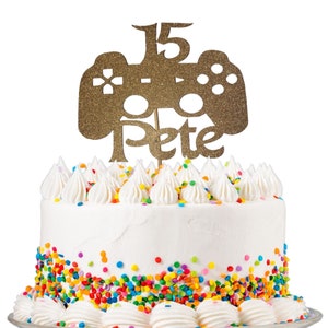 May include: A white cake with colorful sprinkles and a gold glitter cake topper that says "15 Pete" and features a video game controller.
