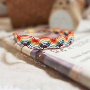 May include: A rainbow colored friendship bracelet with a wavy pattern. The bracelet is made with colorful embroidery floss and is lying on a book with the text "for every"