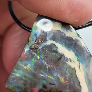 May include: Triangular opal pendant with a black leather cord. The opal features a mix of colors, including green, blue, and red, with a white and brown pattern. The pendant is held by a person's fingers.