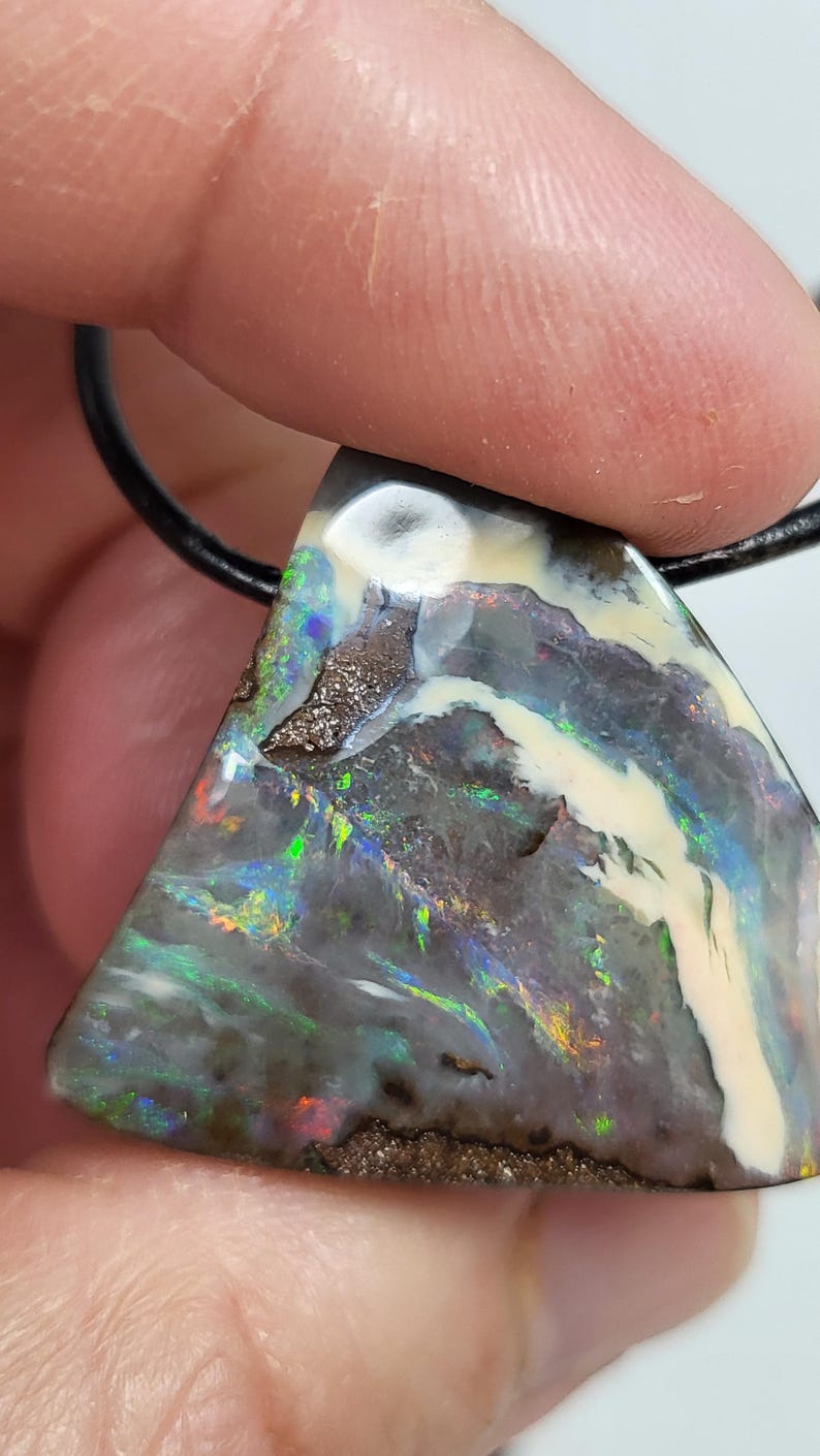 May include: Triangular opal pendant with a black leather cord. The opal features a mix of colors, including green, blue, and red, with a white and brown pattern. The pendant is held by a person's fingers.
