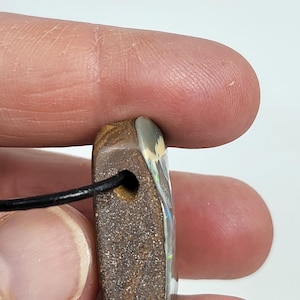 May include: A rectangular pendant with a black cord. The pendant is a mix of brown and tan with a polished, iridescent surface. The pendant has a small hole for the cord to pass through.