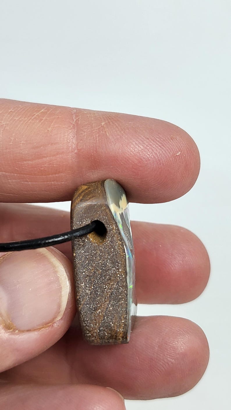 May include: A rectangular pendant with a black cord. The pendant is a mix of brown and tan with a polished, iridescent surface. The pendant has a small hole for the cord to pass through.