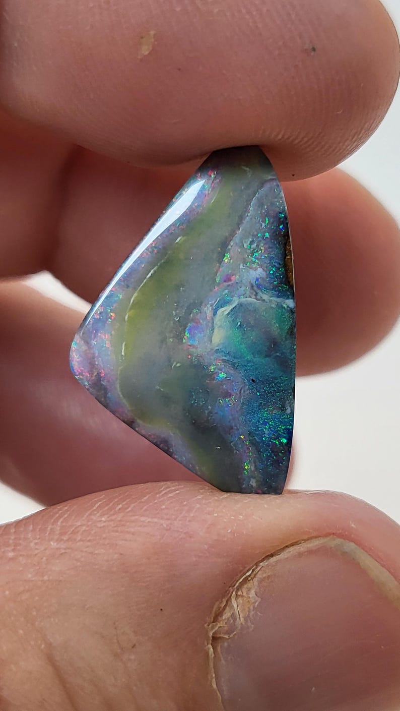 May include: A triangular piece of opal with a dark blue, green, and purple iridescent sheen. The opal has a light green and white pattern.