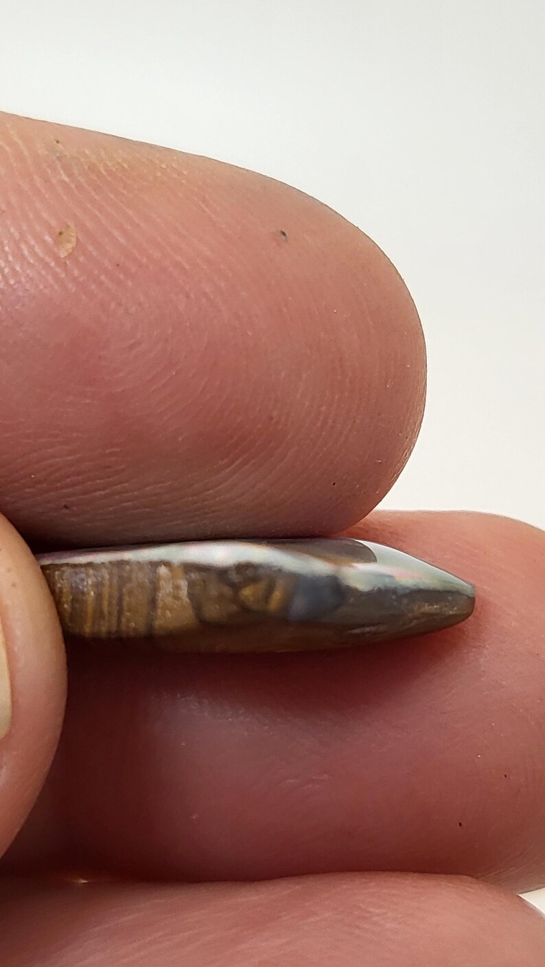 May include: A close-up of a small, teardrop-shaped opal cabochon with brown, white, and gray banding. The opal is set in a silver setting and is being held by a finger.