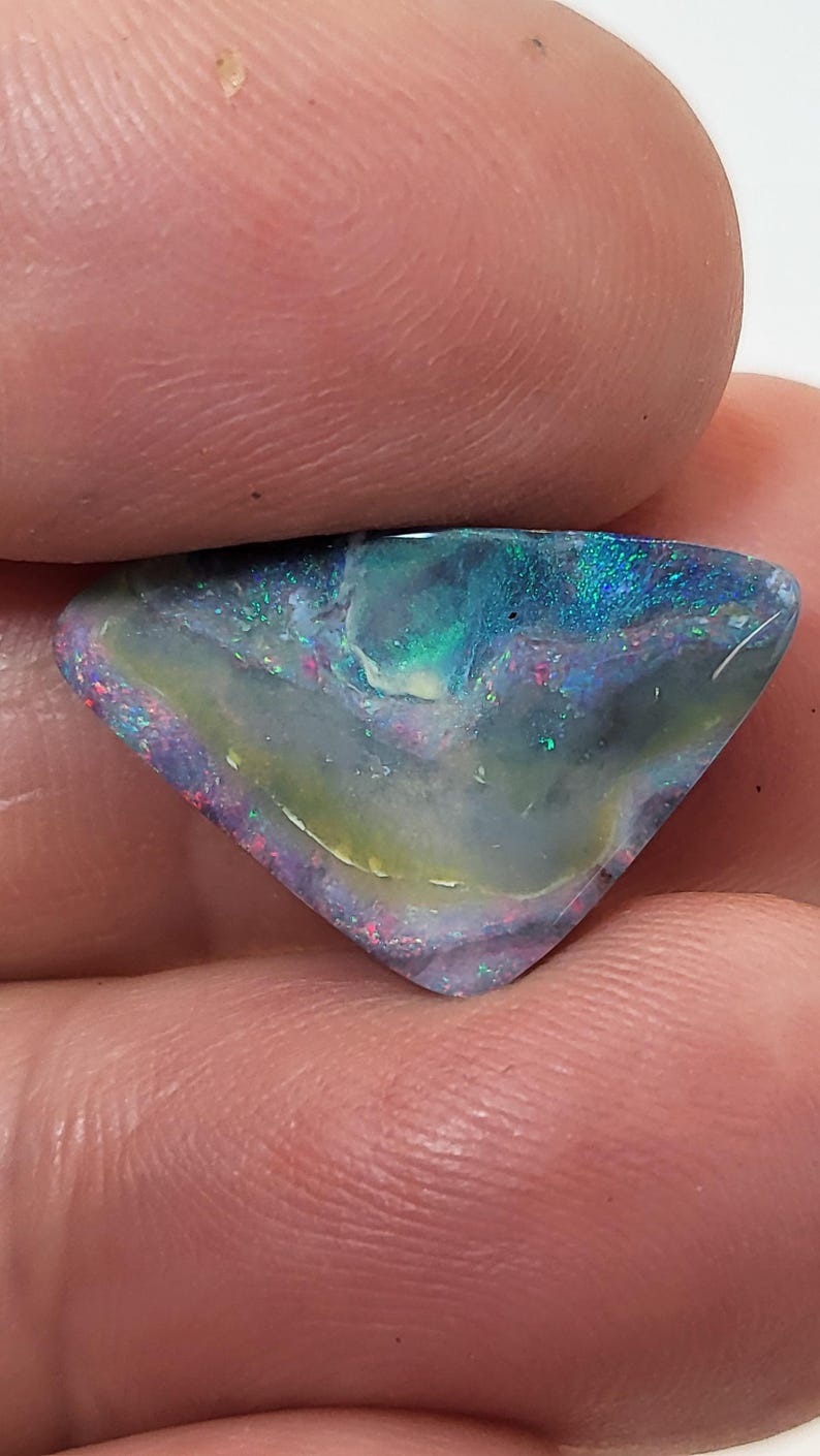 May include: A triangular shaped opal gemstone with a blue, green, and purple iridescent sheen. The opal has a white and yellow base color.