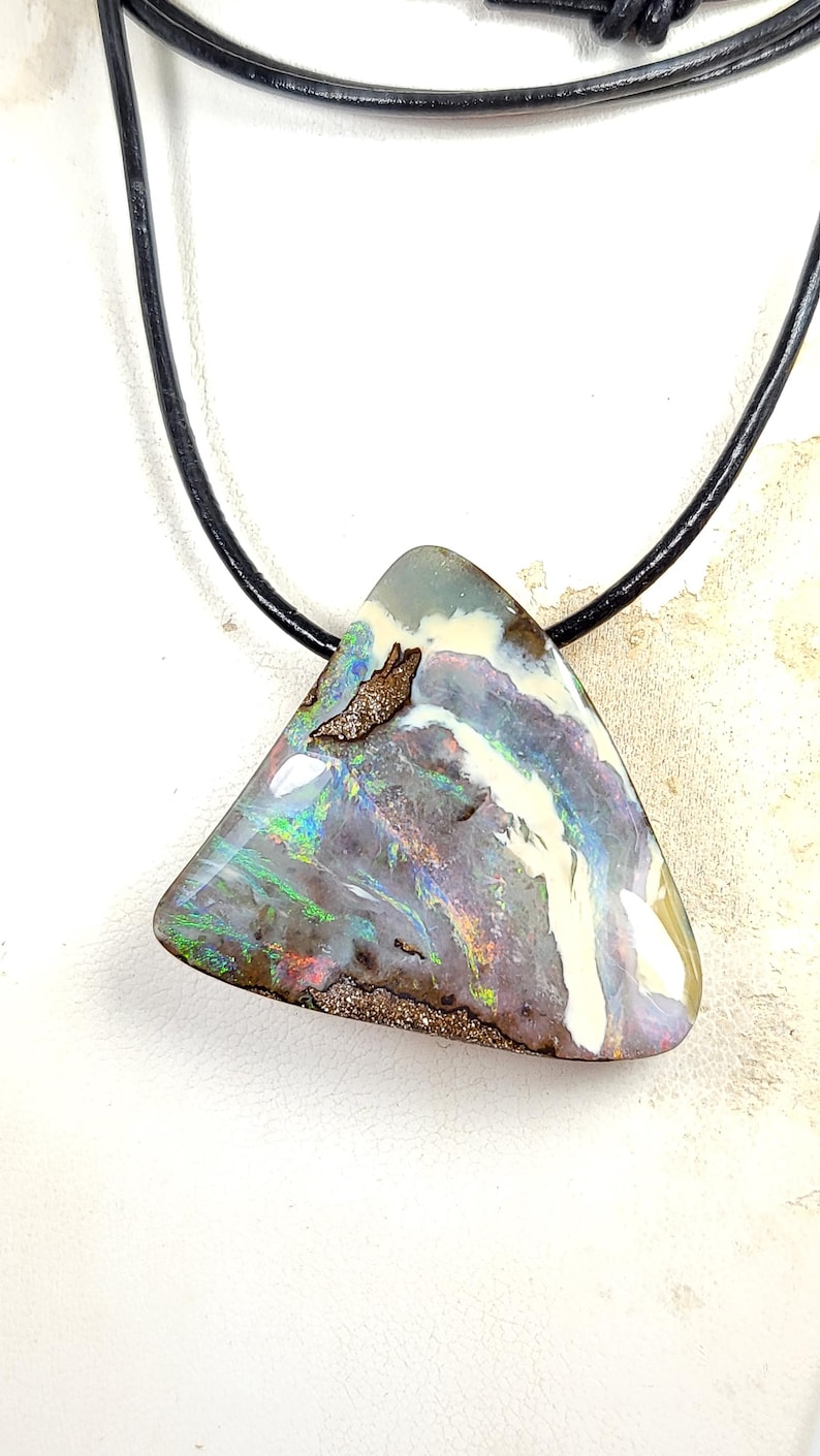 May include: A triangular opal pendant with a black leather cord. The opal features a mix of colors, including white, purple, green, and brown. The pendant is approximately 2 inches wide and 2 inches tall.