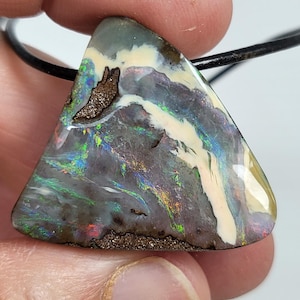 May include: A triangular opal pendant with a black leather cord. The opal features a mix of colors, including green, blue, and red, with a dark gray base and brown matrix. The stone has a polished surface.