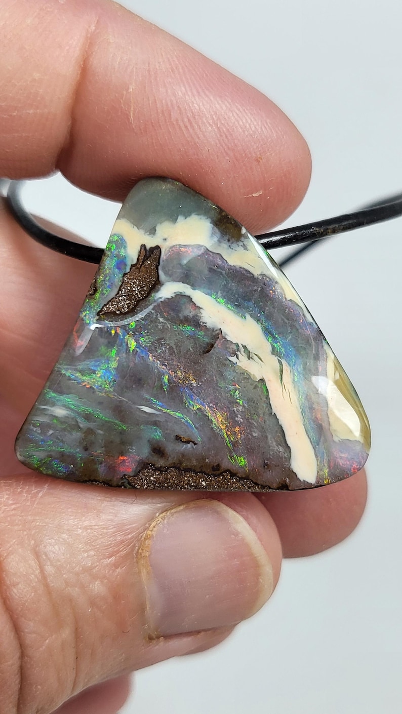 May include: A triangular opal pendant with a black leather cord. The opal features a mix of colors, including green, blue, and red, with a dark gray base and brown matrix. The stone has a polished surface.