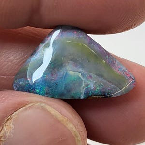 May include: A triangular piece of opal with a blue, green, and white iridescent sheen. The opal has a dark gray base color.