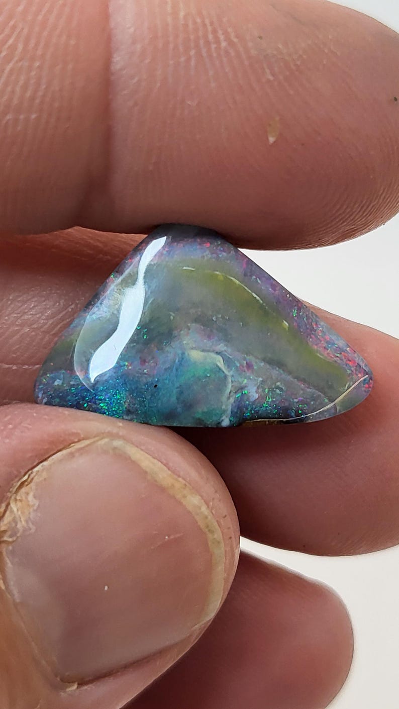May include: A triangular piece of opal with a blue, green, and white iridescent sheen. The opal has a dark gray base color.