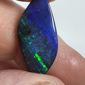 May include: A polished marquise-shaped black opal gemstone featuring vibrant flashes of deep blue and bright neon green colour play across its dark surface. The stone has a smooth, reflective finish and exhibits a distinct play of colour pattern.