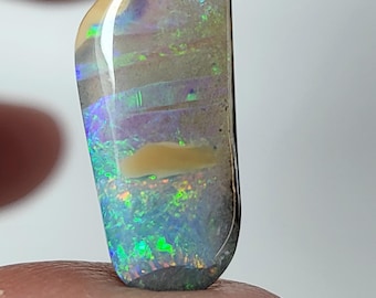 May include: A teardrop-shaped opal gemstone with a dark gray base and iridescent colors, including green, blue, and purple. The opal has a white band across the center.