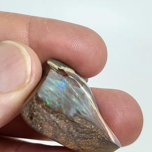 May include: A triangular-shaped opal gemstone with a brown matrix and iridescent play-of-color. The polished surface reflects hues of blue, green, and gold. The stone is held between fingers against a white background.
