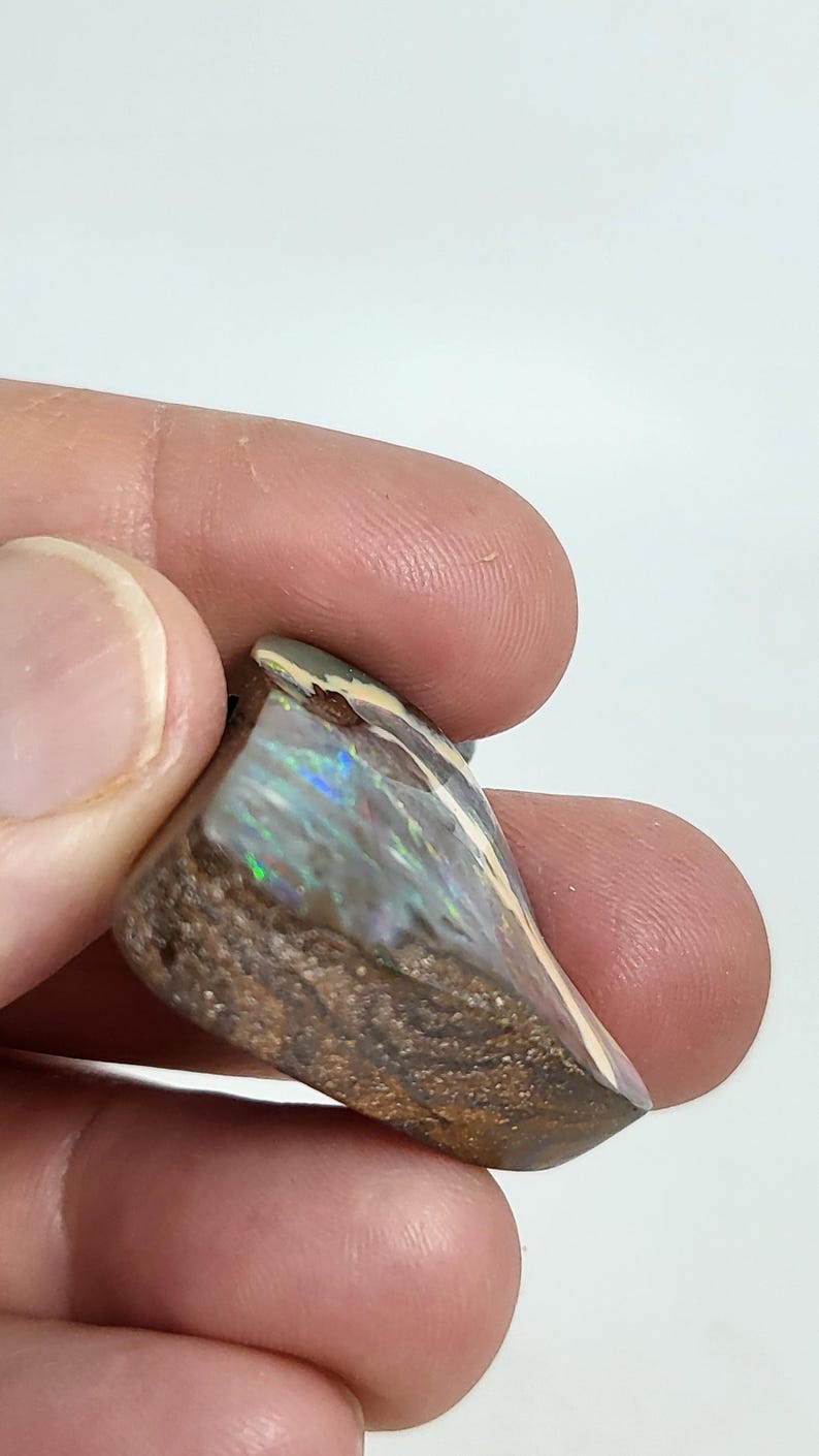 May include: A triangular-shaped opal gemstone with a brown matrix and iridescent play-of-color. The polished surface reflects hues of blue, green, and gold. The stone is held between fingers against a white background.