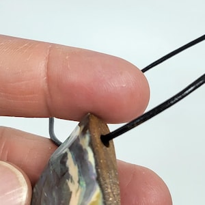 May include: A triangular pendant with a natural opal stone. The stone has a mix of colors, including blue, yellow, and white, and is attached to a black cord. The pendant is held by a person's fingers.