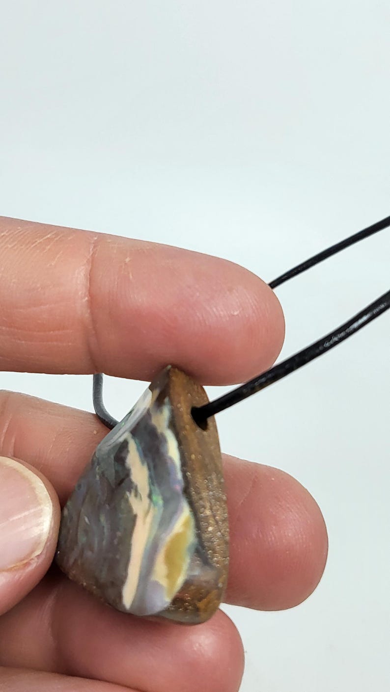 May include: A triangular pendant with a natural opal stone. The stone has a mix of colors, including blue, yellow, and white, and is attached to a black cord. The pendant is held by a person's fingers.