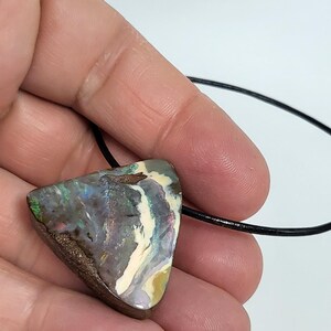 May include: A triangular pendant featuring a vibrant opal stone with a mix of colors including green, blue, and white. The opal is set on a brown base and strung on a black cord, creating a unique and eye-catching necklace.