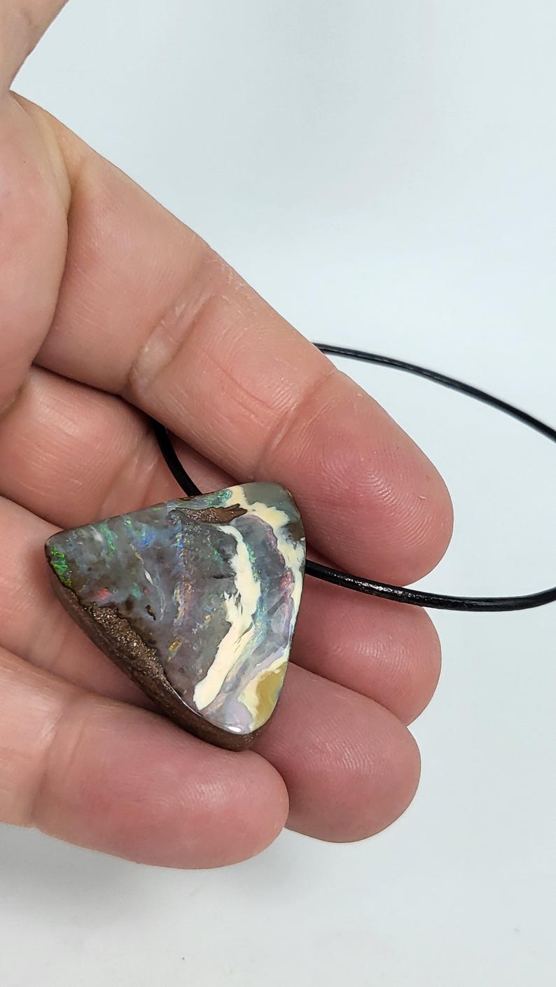 May include: A triangular pendant featuring a vibrant opal stone with a mix of colors including green, blue, and white. The opal is set on a brown base and strung on a black cord, creating a unique and eye-catching necklace.