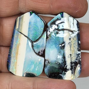 Könnte beinhalten: Zwei irisierende Opal-Cabochons mit einem blauen, grünen und weißen Muster. Die Opale sind in einer silbernen Fassung gefasst und haben eine polierte Oberfläche.