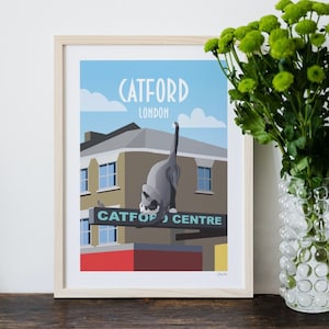 May include: A framed illustration of a cat in front of a building with the text "Catford London" and "Catford Centre" on the sign. The illustration is in a cartoon style with a blue sky and white clouds.