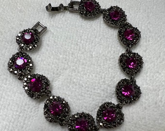 May include: A black bracelet with a silver clasp and purple rhinestones. The bracelet has a total of 10 rhinestones, each set in a circular frame.