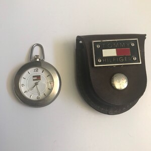 May include: A silver pocket watch with a white face and black hands. The watch is in a brown leather case with a silver Tommy Hilfiger logo.