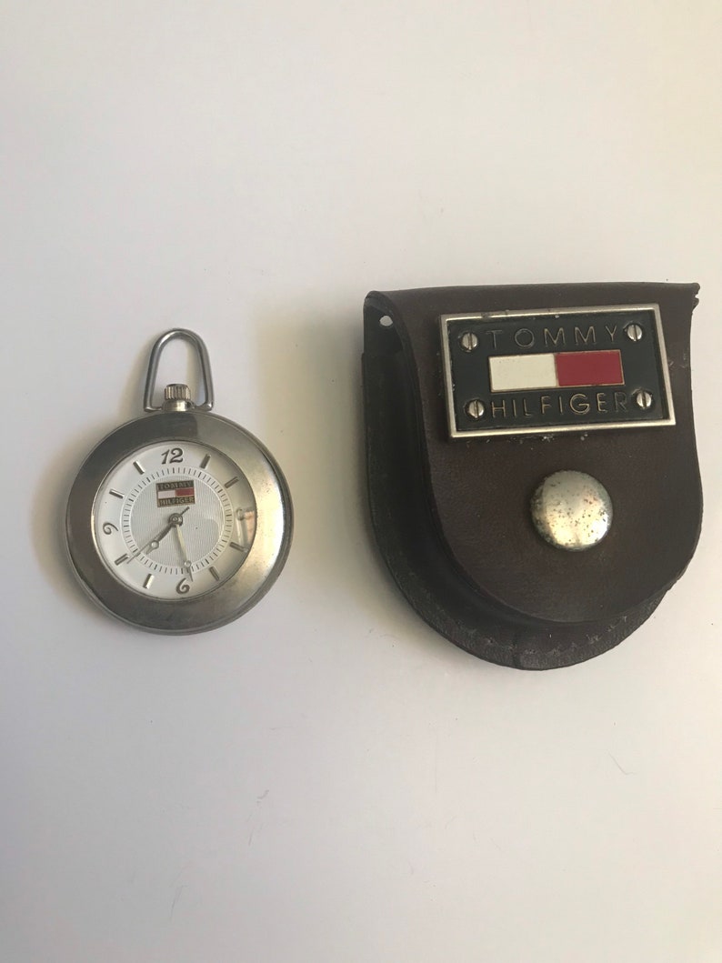 May include: A silver pocket watch with a white face and black hands. The watch is in a brown leather case with a silver Tommy Hilfiger logo.