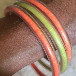 Könnte beinhalten: Zwei durchsichtige Plastikarmbänder mit Glitzer. Ein Armband ist orange und das andere grün.