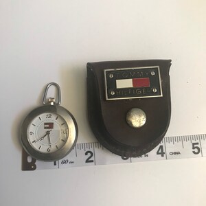May include: A silver pocket watch with a white face and black hands. The watch has a Tommy Hilfiger logo on the face and comes with a brown leather case. The case has a silver Tommy Hilfiger logo with a red, white, and blue flag.