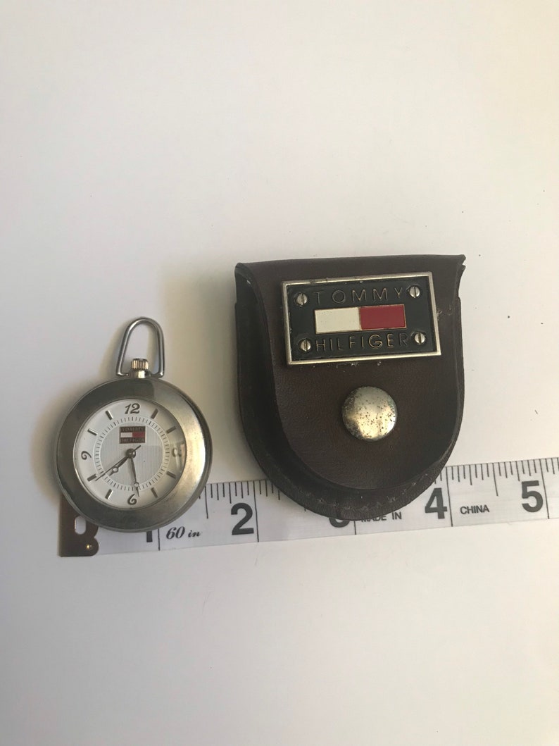 May include: A silver pocket watch with a white face and black hands. The watch has a Tommy Hilfiger logo on the face and comes with a brown leather case. The case has a silver Tommy Hilfiger logo with a red, white, and blue flag.